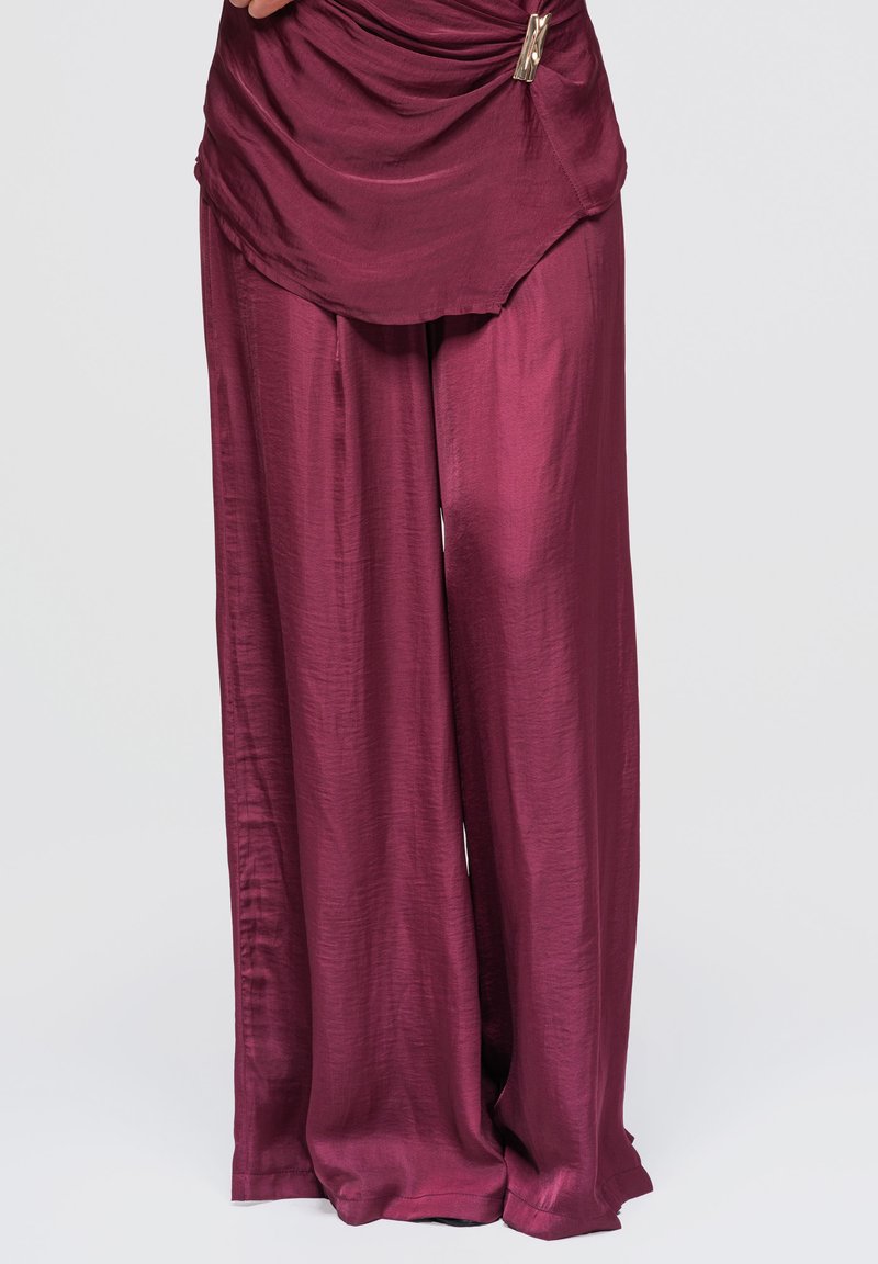 Femme portant un pantalon large bordeaux brillant et ample, ainsi qu'un haut drapé assorti avec un clip métallique sur fond uni.
