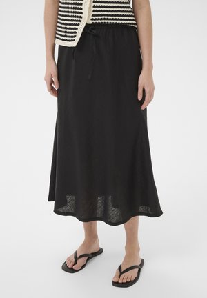 Femme portant une jupe midi noire avec une taille à cordon, un haut rayé noir et blanc, et des sandales noires type tongs sur fond blanc.