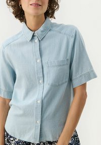 Light blue short-sleeve button-up shirt in denim fabric with a pocket on the left side and a classic collar. Smooth texture.