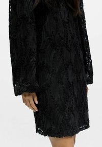 Schwarzes Kleid aus strukturiertem Stoff mit erhabenen, gewellten Mustern. Lange Ärmel und ein leicht ausgefranster Saum verleihen dem Design Detail.