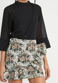 Haut noir transparent à manches longues avec un col haut, associé à une mini-jupe blanche à motifs floraux avec des volants et un détail de nœud sur le côté.