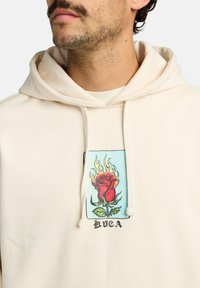 Kremowa bluza z kapturem z grafiką z przodu przedstawiającą czerwoną różę otoczoną płomieniami. Zawiera sznurki oraz nadrukowany tekst "BUCA" poniżej wzoru.