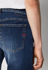 Jeans de denim en azul oscuro con una textura suave. Presenta un logo bordado en rojo en el bolsillo trasero, con costuras naranjas en contraste.