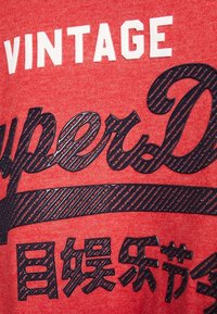 Czerwony materiał z wypukłym czarnym i białym napisem "VINTAGE SuperDry" oraz czarnymi japońskimi znakami poniżej.