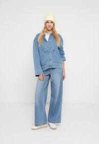 Ljusblå jeansjacka med två framfickor, knappstängning och lös passform, kombinerad med vida jeansbyxor och vita sneakers.