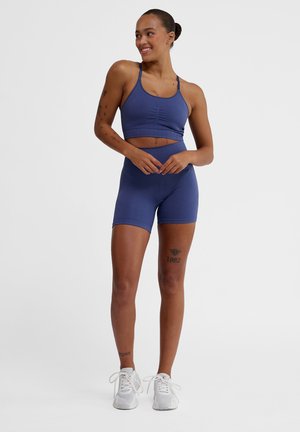 Purple athletic crop top with thin straps and matching high-waisted shorts. Comfy fabric with a textured waistband. White sneakers.