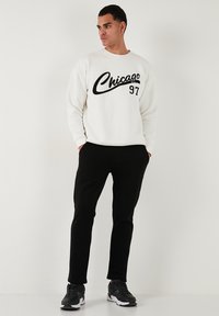 Weißes Sweatshirt mit schwarzem "Chicago 97" Druck, ausgestattet mit Rundhalsausschnitt und langen Ärmeln, kombiniert mit schwarzen Hosen und grauen Sneakern.