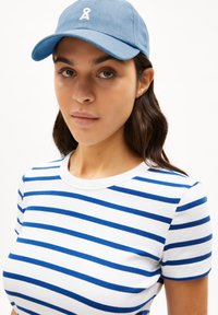 Casquette de baseball bleue avec un logo blanc, associée à un t-shirt blanc avec des rayures horizontales bleues. Gros plan sur le haut du corps et le visage.