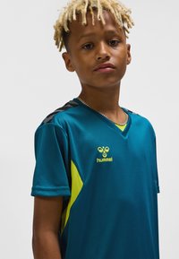 Teal sportströja med korta ärmar, med en gul accent och logotyp. Slät tyg med en v-ringning och svarta detaljer på axlarna.