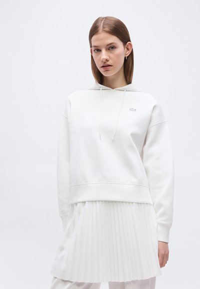Jeune femme portant un sweat à capuche blanc avec cordons et un petit logo, associé à une jupe plissée blanche, sur un fond uni.