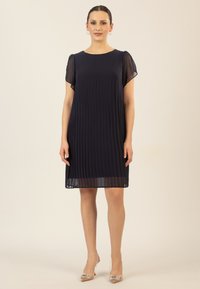Navyblaues Faltenkleid mit transparenten kurzen Ärmeln und rundem Ausschnitt, das oberhalb des Knies endet. Kombiniert mit transparenten spitzen High Heels.