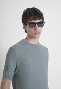 Gafas de sol triangulares negras con lentes oscuros. Llevadas con una camisa de punto de manga corta de color azul claro que presenta un patrón texturizado.