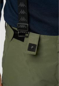 Olive zöld nadrágok letisztult dizájnnal rendelkeznek, fekete állítható vállpántokkal, textúrázott derékpánttal és gombos záródással.