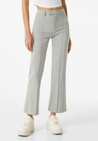 Kobieta w jasnoszarych, dopasowanych spodniach w prostym kroju i białych sportowych butach na platformie, stojąca na tle jednolitej białej ściany.