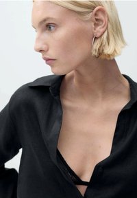 Černá lněná košile s částečně rozepnutým límečkem, pod kterým se objevuje černá krajková podprsenka. Modelka má krátké blond vlasy a stříbrné kruhové náušnice.