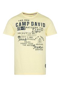 Camiseta de algodón amarilla con gráficos de texto mixto en azul marino y blanco, cuello redondo, mangas cortas y un pequeño parche de logo en el costado.