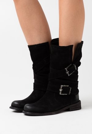 Botas de ante negras hasta la mitad de la pantorrilla con detalles de correa y hebilla plateada, usadas sobre piernas desnudas, con fondo blanco.