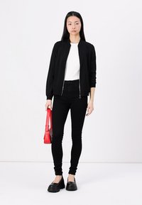 Veste aviateur noire avec fermeture éclair, chemise blanche ajustée, jean skinny noir et grosses chaussures noires ; tenant un sac à main rouge. Fond blanc.