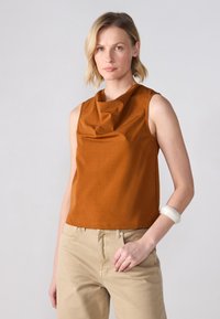 Ærmeløs top lavet af glat, varmtonet stof med en draperet halsudskæring, parret med lysebeige bukser og et uigennemsigtigt, rundt armbånd.