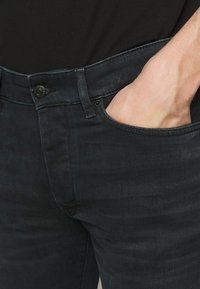Jeans de mezclilla oscura con un corte ajustado, que cuentan con un diseño de bolsillo visible y costuras tonales. La tela tiene una textura suave con un ligero desgaste.