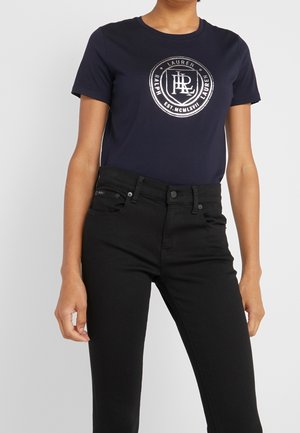 Person iført en mørkeblå Ralph Lauren t-shirt med sølvlogo og sorte højtaljede jeans mod en ensfarvet baggrund.