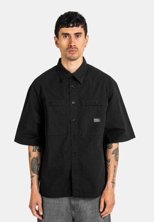 Mann mit kurzen dunklen Haaren, der ein schwarzes kurzärmeliges Hemd und graue Hose trägt, steht vor einem schlichten hellen Hintergrund.