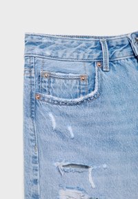 Jeans de mezclilla azul claro con detalles desgastados, que cuentan con bordes deshilachados, un pequeño bolsillo y remaches de metal en la cintura.