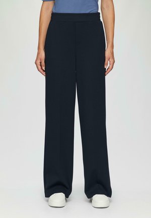 Marineblaue, weit geschnittene Hose aus glattem Material mit elastischem Bund und vorderen Nähten. Kombiniert mit weißen Sneakern, was einen lässigen Look ergibt.