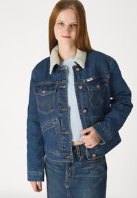 Τζάκετ από τζιν με ελαφρώς μπεζ γούνινο γιακά, μπλε ύφασμα, μπροστινές τσέπες και χάλκινους κουμπαράδες. Φοριέται πάνω από ανοιχτό μπλε μπλουζάκι.