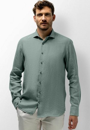 Mann mit kurzen braunen Haaren und Bart, der ein langärmeliges hellgrünes Hemd und hellbeige Hose trägt, steht vor einem weißen Hintergrund.