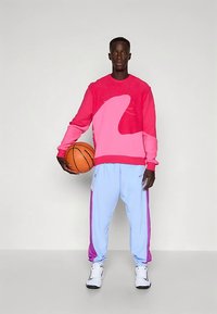 Svetlo roza pulover z valovitim vzorcem, v kombinaciji s svetlo modrimi trenirkami z roza poudarki. Model drži košarkarsko žogo.