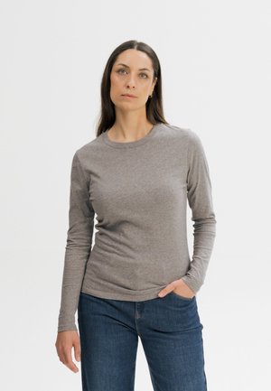 Vrouw met lang donker haar, die een grijs shirt met lange mouwen en blauwe spijkerbroek draagt, staand met één hand in haar zak tegen een effen achtergrond.