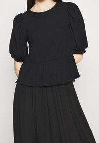 Černá blůza s nafouknutými rukávy, texturovaným povrchem a peplum pasem. Výstřih je kulatý bez dalšího zdobení.