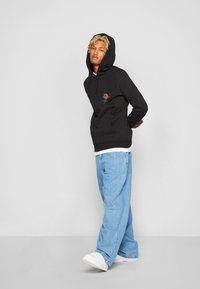 Black hoodie with rose embroidery on the front, paired with light blue baggy jeans and white sneakers. Simple design and casual fit.