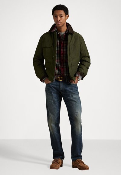 Chaqueta verde oliva con cuello marrón sobre una camisa de cuadros rojos, combinada con vaqueros azules y zapatos marrones, de pie contra un fondo blanco.