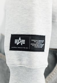 Étiquette noire Alpha Industries avec logo et texte blanc sur bord de manche en tissu gris clair.