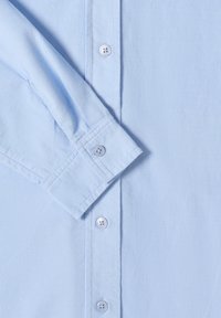 Chemise bleu clair à manches longues avec poignets boutonnés visibles et trois boutons en plastique transparent sur la patte de boutonnage avant.