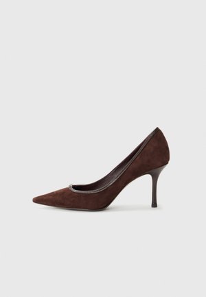 Escarpin en suède marron avec un bout pointu, une texture lisse et un talon fin. Présente un accent métallique autour de l'ouverture.