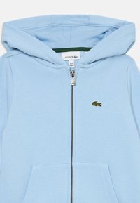 Svijetloplava majica s patentnim zatvaračem izrađena od mekog materijala, s džepom za kengure, kapuljačom i izvezenim zelenim Lacoste logotipom.