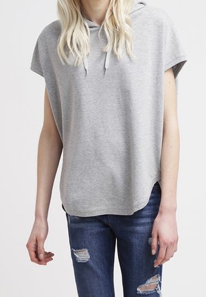 Persoon die een lichtgrijze mouwloze hoodie met trekkoorden draagt en gescheurde donkerblauwe spijkerbroek, staand tegen een effen witte achtergrond.