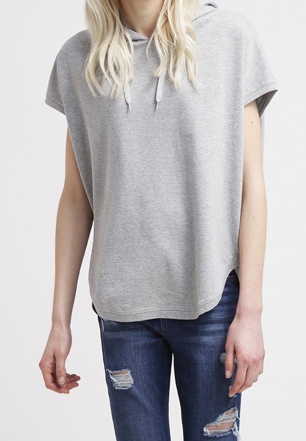 Personne portant un sweat à capuche sans manches gris clair avec cordons et un jean bleu foncé déchiré, debout devant un fond blanc uni.