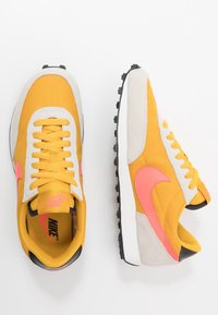 Gula sneakers med gråmocka detaljer, rosa Nike-swoosh, vit mellansula och svart gummisula. Utrustad med snörning och vadderad krage.