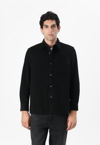 Chaqueta de pana negra con mangas largas, cierre de botones, un bolsillo en el pecho y un acabado texturizado. Combinada con jeans oscuros.