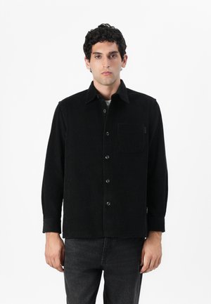 Black corduroy jacket with long sleeves, button closure, single chest pocket, and a textured finish. Paired with dark jeans.
