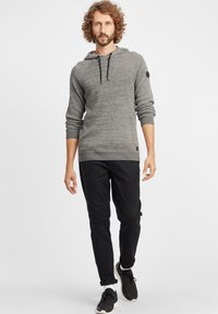 Szary dzianinowy hoodie z sznurkiem, ściągaczami i teksturowanym wzorem. Noszony z czarnymi spodniami i czarnymi trampkami.
