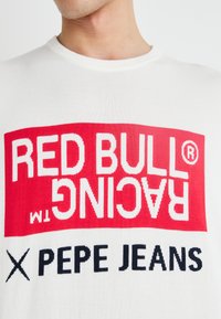 Fehér pamut póló, piros grafikával, amelyen a "RED BULL RACING" fehér szöveggel és az "X PEPE JEANS" fekete szöveggel található a mellkas területén.