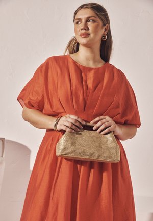 Piros lenruha laza ujjas, redőzött kialakítással és egy kis arany szőtt clutch táskával. Egyszerű arany ékszerek kísérik.