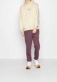Felpa beige con cappuccio, tasca frontale e logo, abbinata a pantaloni jogger viola. Le calzature presentano dettagli bianchi, arancioni e grigi.