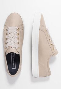 Beige Canvas-Sneaker in niedriger Ausführung mit weißen Gummisohlen, goldfarbenen Ösen, gestreiften Schnürsenkeln und Tommy Hilfiger-Logo auf der Zunge und der Einlegesohle.