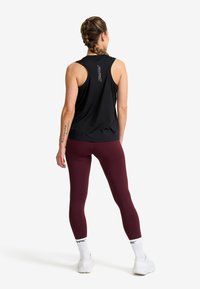 Woman with braided hair stands facing away wearing a black sleeveless top, maroon leggings, white socks, and white sneakers on white background.